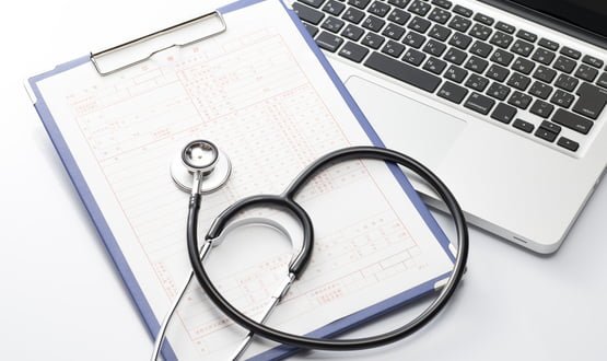 A stethoscope lies on top of a clipboard next to a laptop